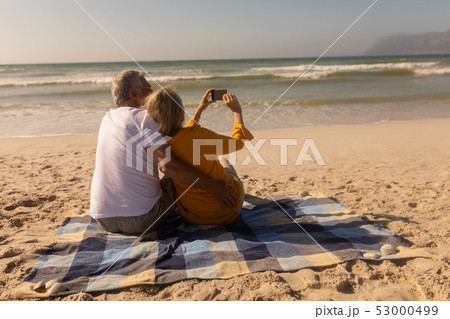 Senior couple taking selfie with mobile phone relaxing on picnic blanket at beach Senior couple taking selfie with mobile phone relaxing on picnic blanket at beach 53000499