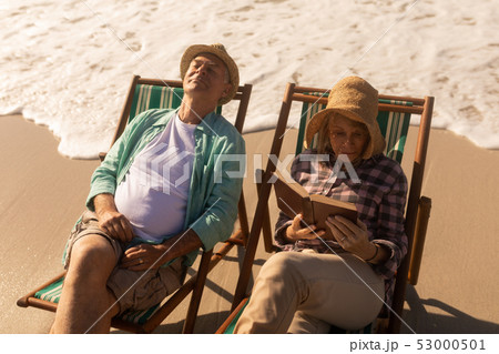 Senior woman reading a book while senior man relaxing on sun lounger at beach 53000501