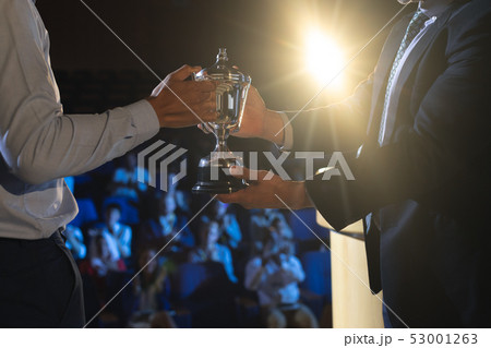 Businessman giving trophy to business male executive on stage in auditorium Businessman giving trophy to business male executive on stage in auditorium 53001263