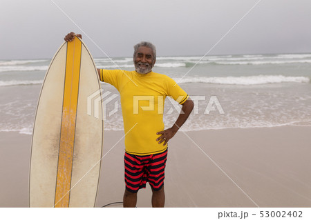 Senior male surfer standing with surfboard and looking at camera on the beach  53002402