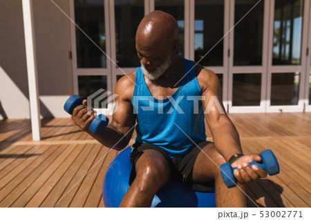 Senior man balancing on exercise ball while training biceps with dumbbells in the backyard of home 53002771