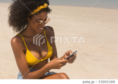 Young African American woman in yellow bikini and sunglasses using mobile phone at beach 53003166