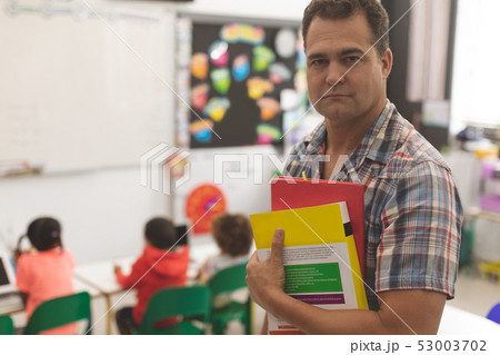 Schoolteacher standingin classroom with book and looking at the camera at school Schoolteacher standingin classroom with book and looking at the camera at school 53003702