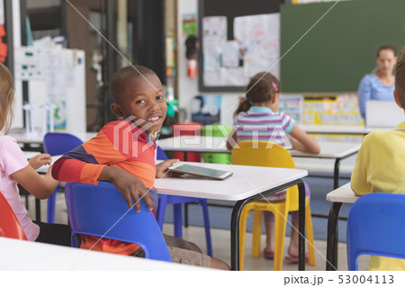Happy schoolboy looking at the camera and holding digital tablet at desk in classroom Happy schoolboy looking at the camera and holding digital tablet at desk in classroom 53004113