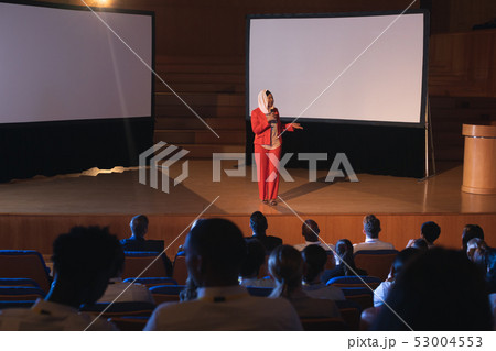 Businesswoman standing and giving presentation in auditorium  53004553