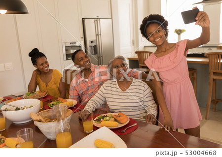 Girl taking selfie of family with mobile phone on dining table at home 53004668
