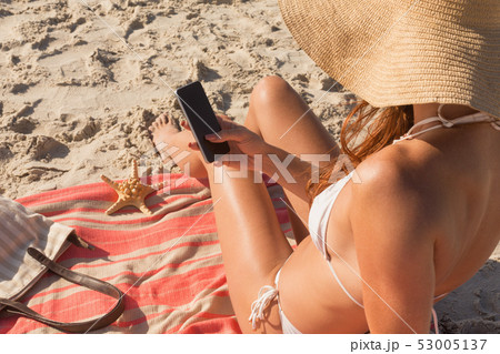 Young Caucasian woman in hat and bikini using her mobile phone on the beach Young Caucasian woman in hat and bikini using her mobile phone on the beach 53005137