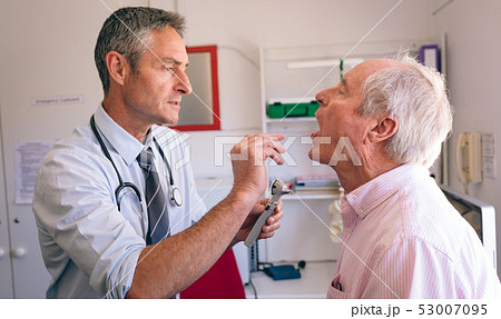 Male doctor checking senior male patient mouth with otoscope 53007095