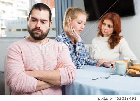 Upset young man sitting at table after quarrel and girl with mother on background Upset young man sitting at table after quarrel and girl with mother on background 53013491