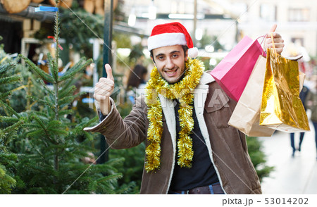Man in Christmas hat with purchases delighted at Christmas fair, thumbs up 53014202