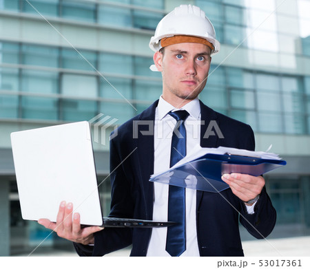 Architector in suit and hat is looking into documents with project and comparing in laptop 53017361
