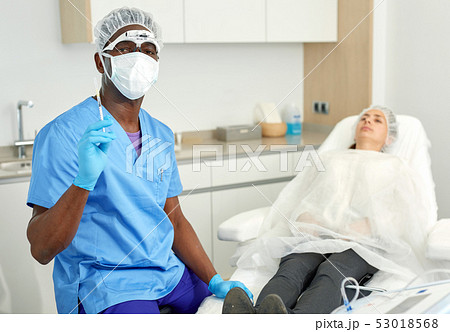 Cosmetician male in mask before aesthetic facial injection, woman in amchair Cosmetician male in mask before aesthetic facial injection, woman in amchair 53018568