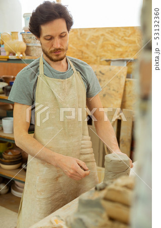 Focused dark-haired man in special apron measuring needed amount of clay 53132360