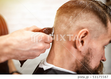 Master cuts hair and beard of men in the barbershop, hairdresser makes hairstyle for a young man 53175587