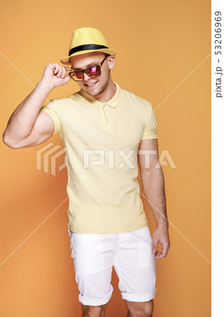 Confident stylish guy in yellow shirt, sunglasses, white shorts, straw hat looking side away from 53206969