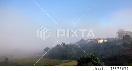 Morning fog over rice fields, sun shines through to small clay buildings - typical landscape in 53378637