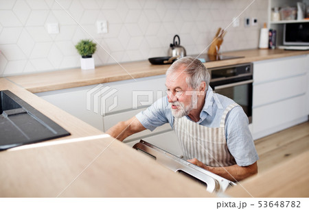 A senior man indoors in kitchen at home, loading dishwasher. 53648782