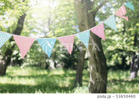 Pastel bunting flags hanging among trees. Summer garden party. Outdoor birthday, wedding decoration Pastel bunting flags hanging among trees. Summer garden party. Outdoor birthday, wedding decoration 53650460
