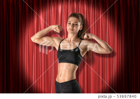 Crop image of young fit woman in crop top, standing in half-turn in spotlight at red stage curtain 53697840