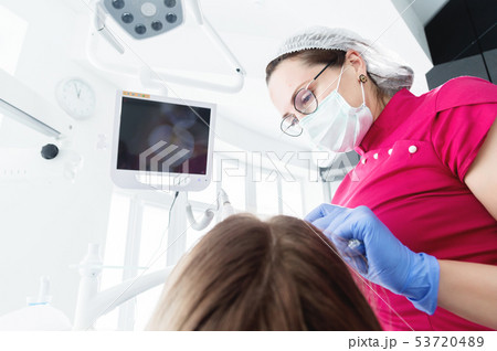 A professional dentist woman in glasses and overalls examines the oral cavity of a young girl in the 53720489