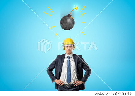Crop image of businessman in yellow hard hat with ear defenders, standing with hands on hips, and 53748349