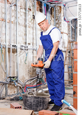 Construction worker mixing dry mortar with handheld mixer in bucket in overhauls indoors Construction worker mixing dry mortar with handheld mixer in bucket in overhauls indoors 53771813