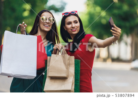 Beautiful happy girls in sun glasses holding shopping bags, making selfie on smart phone and smiling Beautiful happy girls in sun glasses holding shopping bags, making selfie on smart phone and smiling 53807894