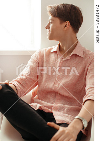 Handsome young man of model appearance is sitting by the window indoor in a shirt and black jeans Handsome young man of model appearance is sitting by the window indoor in a shirt and black jeans 53831472