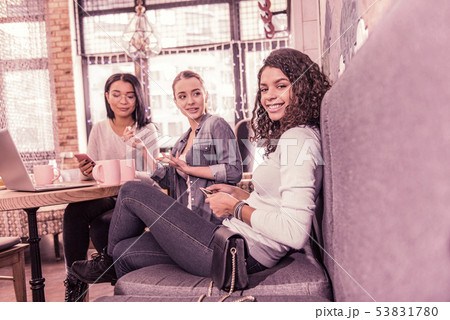 Beaming curly dark-haired woman sitting on grey sofa in cafeteria with her friends 53831780