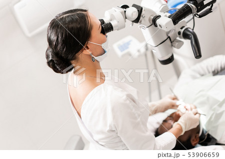 Young woman dentist treating root canals using microscope in the dental clinic. Man patient lying on Young woman dentist treating root canals using microscope in the dental clinic. Man patient lying on 53906659
