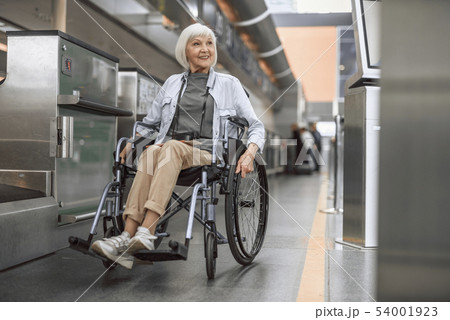 Cheerful mature woman on disabled carriage moving near check-in counter Cheerful mature woman on disabled carriage moving near check-in counter 54001923