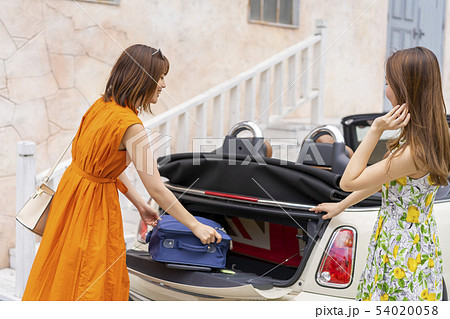 オープンカーのトランクにキャリーバッグを入れ女子旅に出かける可愛いワンピースを着た二人の女性 54020058