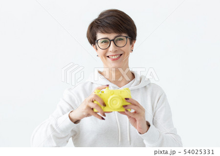 Positive middle-aged woman in glasses holds in hand a vintage yellow camera posing on a white 54025331