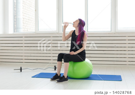 Charming slim young girl hipster with colored hair drinks water while sitting on a fitball next to a 54028768
