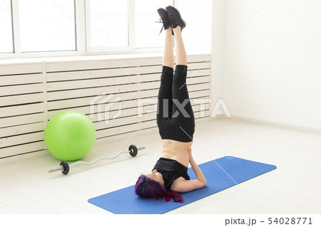 Young slim girl with colored hair makes an headstand on a sports blue mattress next to a barbell and 54028771