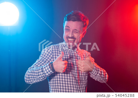 Laughing young bearded man in a plaid shirt showing thumb up posing in studio on blue-red background 54045288