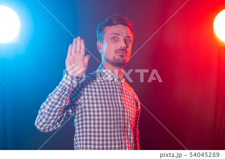 Serious young bearded handsome hipster man shows palm up portraying stop on blue-red background 54045289