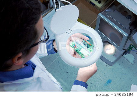 A man in a white robe and glasses checks the electrical Board for defects through a magnifying glass 54085194