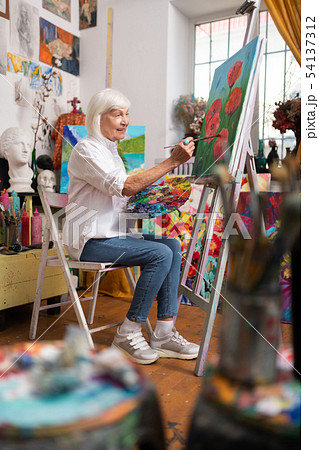 Aged woman wearing jeans sitting and painting on canvas 54137312
