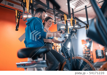 Overweight woman smiling cycling in gym and talking to trainer 54171220
