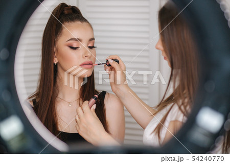 Photo though the ring lamp of makeup artist doing a makeup for attractive young girl. Background of 54240770