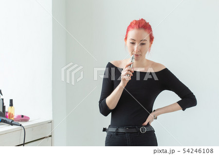 Youth and addiction concept - young red haired woman smoking vape near the window 54246108
