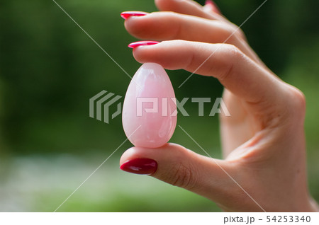 Close up photo of female hand holding a vaginal yoni egg. Rose quartz crystal egg on river 54253340
