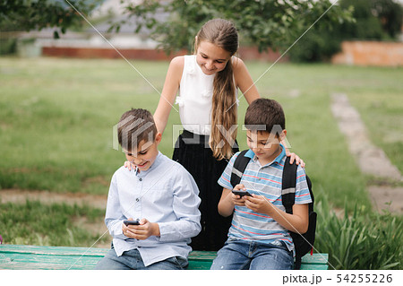 Two boys and girl use their phones during school breack. Cute boys sitting on the bench and play Two boys and girl use their phones during school breack. Cute boys sitting on the bench and play 54255226