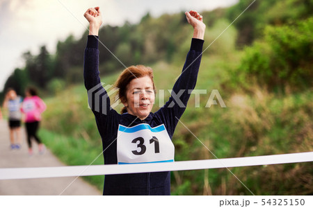 Senior woman runner crossing finish line in a race competition in nature. 54325150