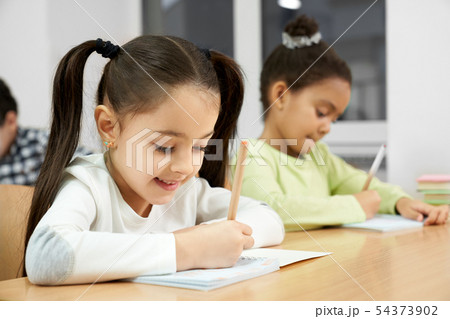 Pretty girls sitting at desk in class, writing in copybook. 54373902
