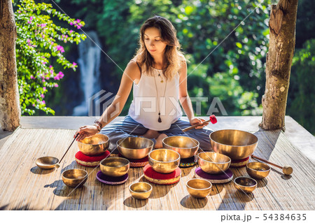 Woman playing on Tibetan singing bowl while sitting on yoga mat against a waterfall. Vintage tonned Woman playing on Tibetan singing bowl while sitting on yoga mat against a waterfall. Vintage tonned 54384635