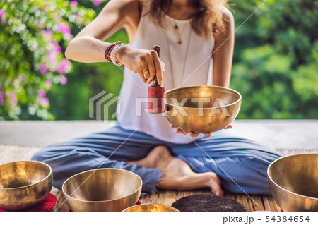 Woman playing on Tibetan singing bowl while sitting on yoga mat against a waterfall. Vintage tonned 54384654