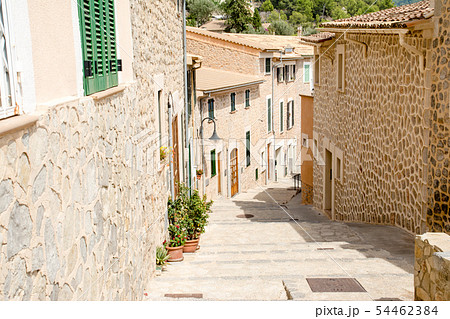 マヨルカ島の街並み　石畳の細道に並ぶ植木と緑の遮光扉の付いた窓のある薄あずき色の壁と石壁の家々 54462384