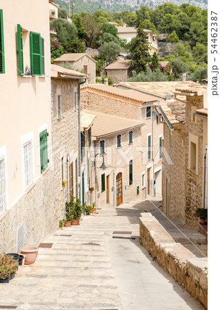 マヨルカ島の街並み　石畳の細道に並ぶ植木と緑の遮光扉の付いた窓のある薄あずき色の壁と石壁の家々 54462387